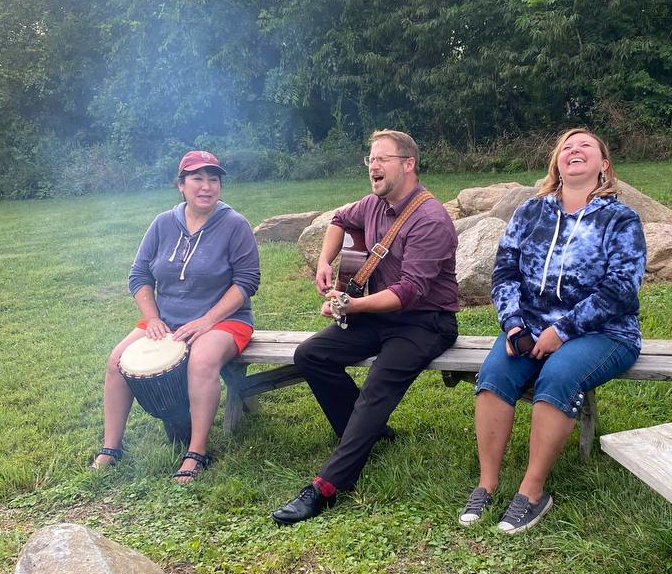 three adults are gathered near a fire playing instruments and laughing